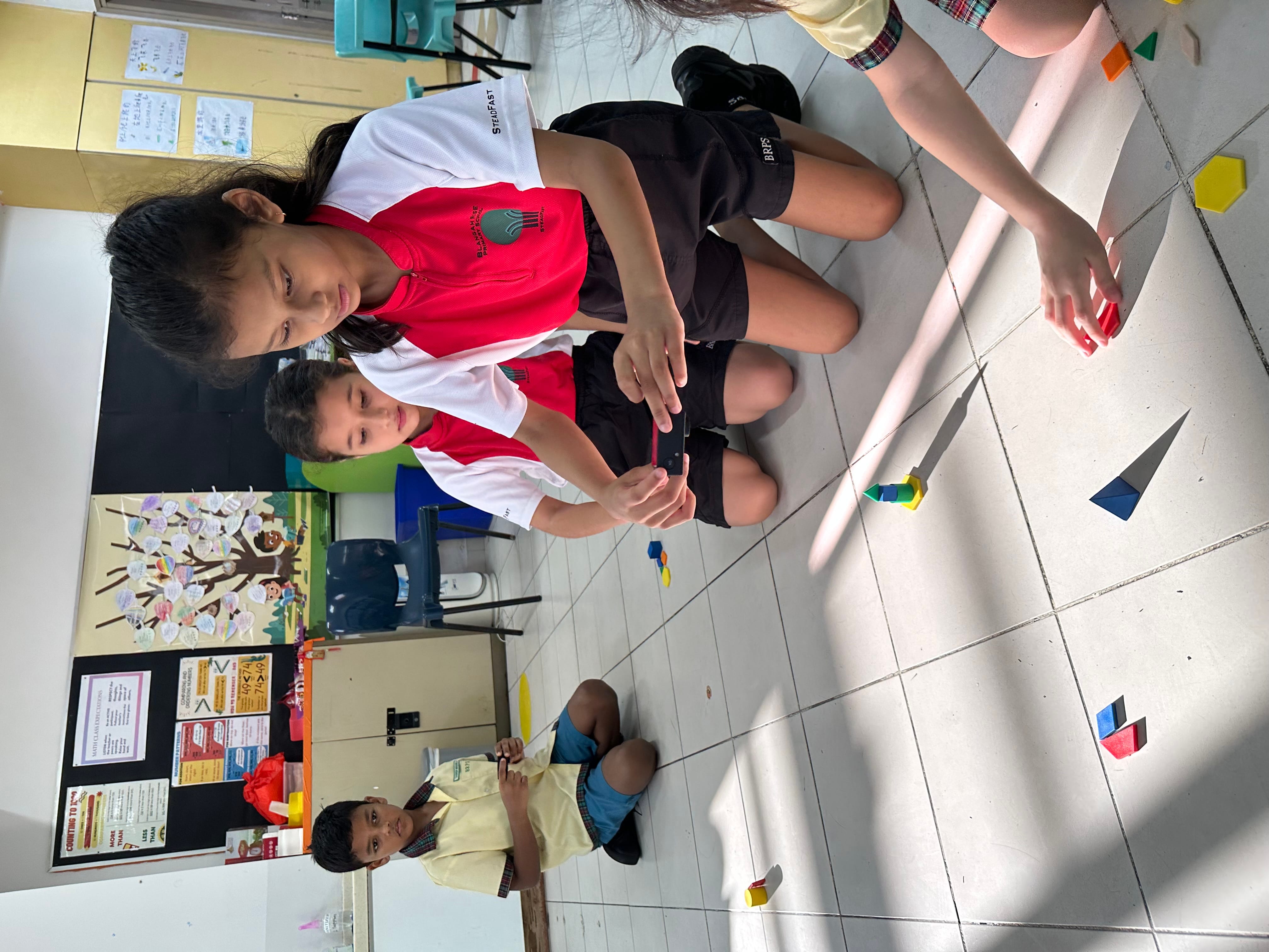 Children in school uniforms sit on a classroom floor arranging colorful wooden geometric shape blocks.